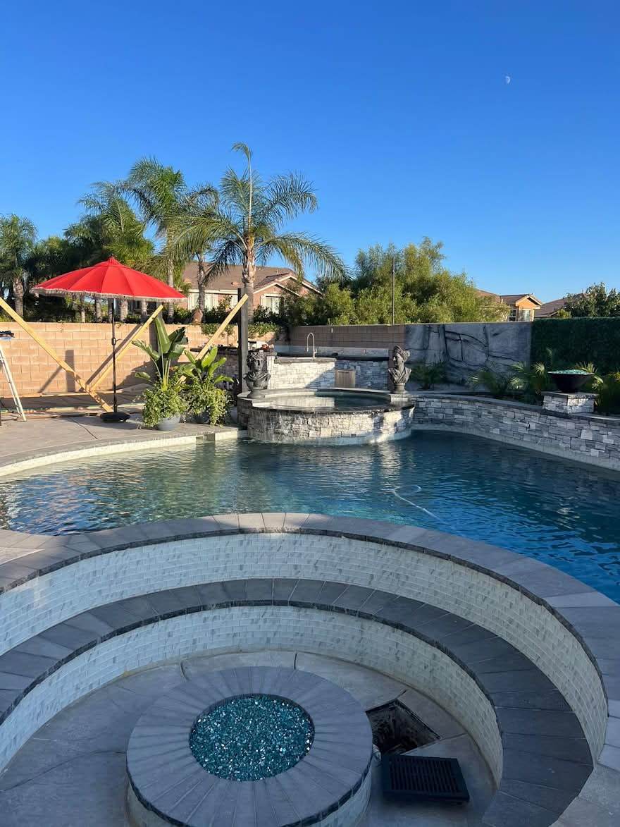 A finished backyard pool with clear blue water, a raised stacked-stone spa spilling into it, statue fountains along the far wall and a sunken firepit filled with teal fire glass in the foreground.