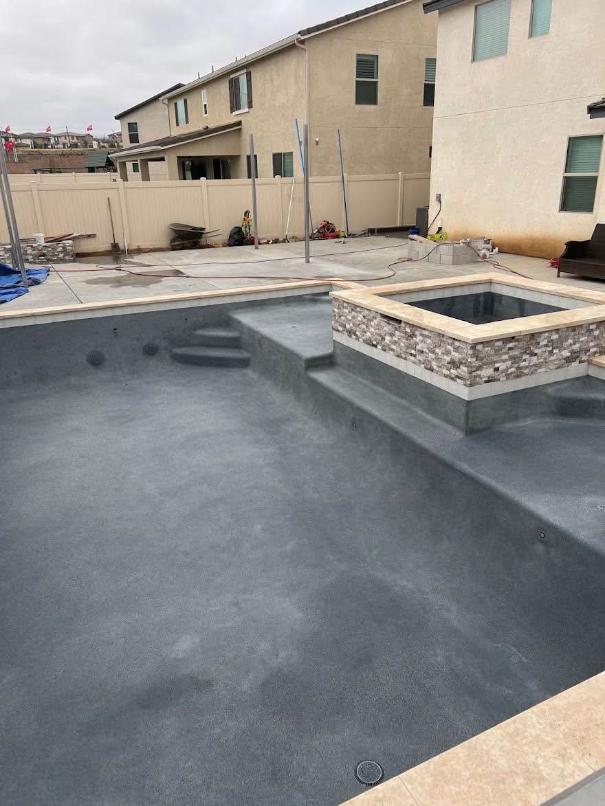 A pool mid-build with its shell freshly coated in smooth grey plaster, a raised square spa with stone veneer at the far end, and the surrounding deck still bare concrete.