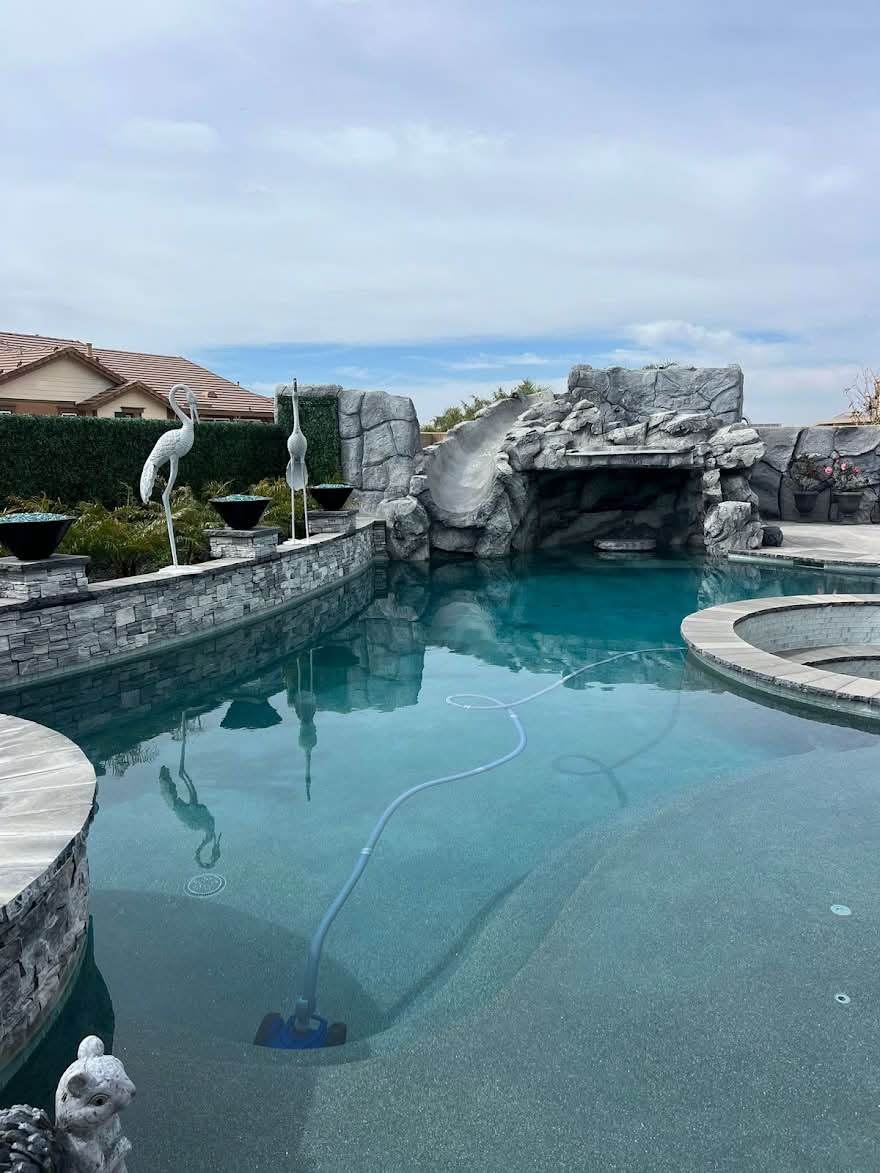 A large backyard pool with a rock grotto and slide at one end, a raised spa, fire bowls along the deck and crane statues at the waterline, filled with clear blue water.