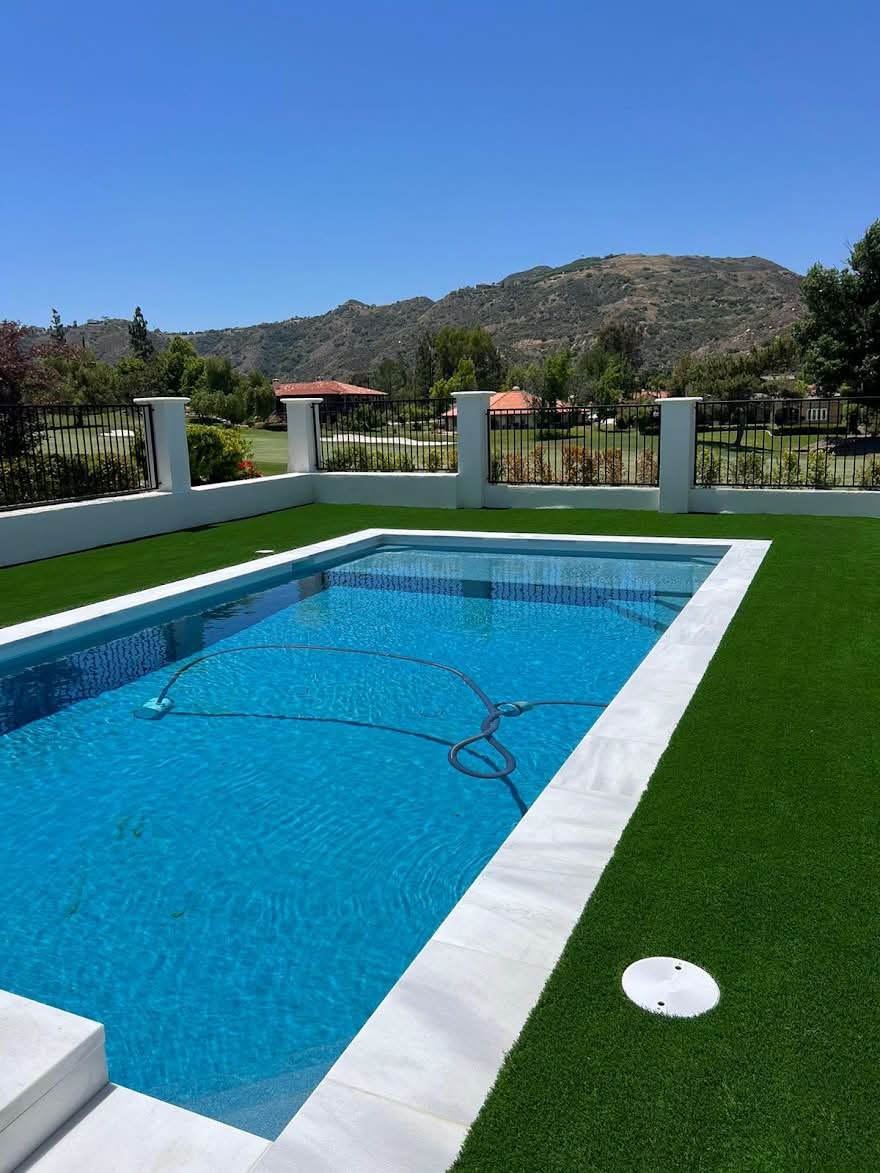 A modern rectangular pool with clear blue water and a wide tiled coping edge, surrounded by turf, looking out over a hillside view.