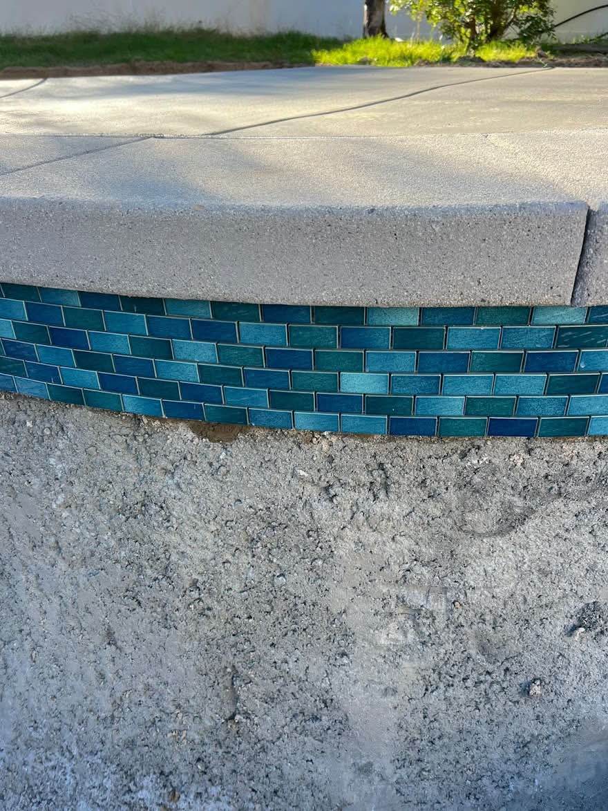 A close-up of a new teal glass waterline tile band being set against the bare grey concrete of a pool wall, with the older rough surface still exposed below it.