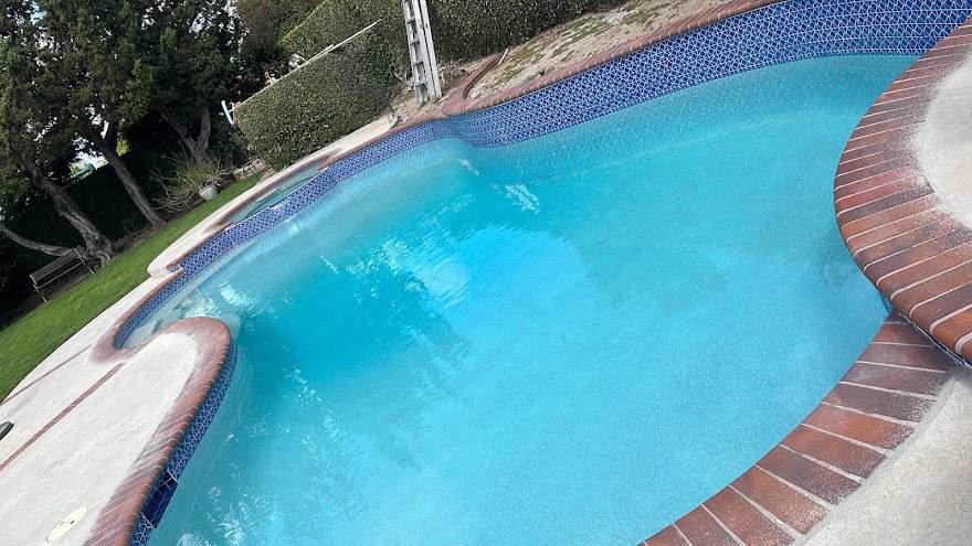 A kidney-shaped pool after resurfacing, its new blue waterline tile band and red brick coping meeting bright turquoise water, with a turf lawn running up to the edge.